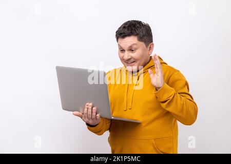 Der Mann winkt mit der Hand und hallo auf den Laptop-Bildschirm, spricht mit einem Kollegen in einem Videogespräch, kommuniziert über eine Online-Konferenz und trägt einen Hoodie im urbanen Stil. Innenaufnahme des Studios isoliert auf weißem Hintergrund. Stockfoto