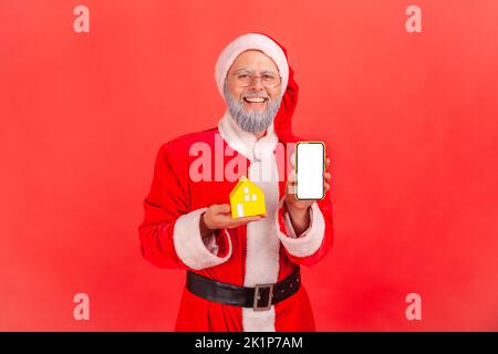 Porträt eines lächelnden älteren Mannes mit grauem Bart, der ein weihnachtsmann-Kostüm trägt und ein kleines Papierhaus und ein Smartphone mit leerem Bildschirm hält. Innenaufnahme des Studios isoliert auf rotem Hintergrund. Stockfoto