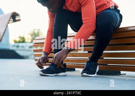 Junger Athlet oder Läufer in roter Sportjacke und schwarzen Leggins, die auf einer Holzbank sitzen und Schuhe von Sneakern binden Stockfoto