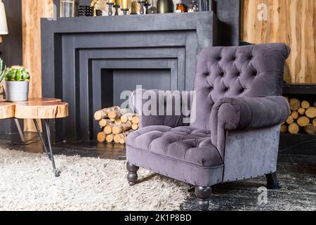 Graublauer Sessel in der Nähe von Holztisch im Wohnzimmer mit Kamin. Fragment des Innenraums eines gemütlichen Hauses. Ein grauer Sessel von einem Stockfoto