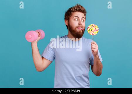 Porträt von lustig gut aussehenden bärtigen Mann mit rosa Hantel ausarbeiten und Blick auf leckere Lollypop mit der Zunge heraus, will Süßigkeiten beißen. Innenaufnahme des Studios isoliert auf blauem Hintergrund. Stockfoto