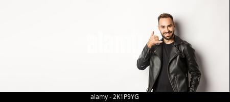 Schöner Mann in Lederjacke flirtet mit Ihnen, zeigt Handy rufen Sie mich Geste, vor weißem Hintergrund stehen Stockfoto