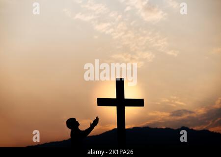 Silhouette eines Mannes Gebet vor dem Kreuz auf dem Berg bei Sonnenuntergang. Begriff der Religion. Stockfoto