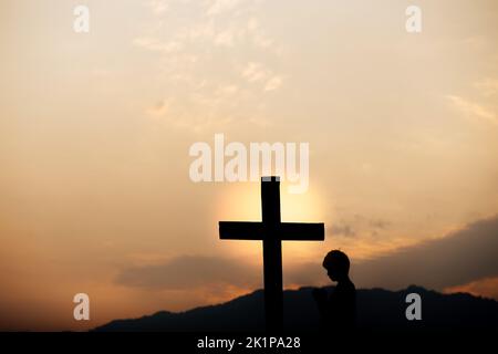 Silhouette eines Mannes Gebet vor dem Kreuz auf dem Berg bei Sonnenuntergang. Begriff der Religion. Stockfoto