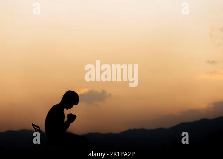 Silhouette eines Mannes, der die Hände hält und am Berg bei Sonnenuntergang betet. Begriff der Religion. Stockfoto