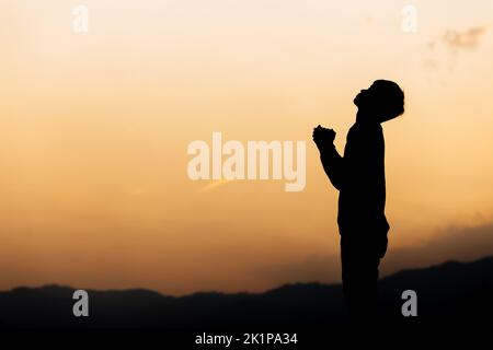 Silhouette eines Mannes, der die Hände hält und am Berg bei Sonnenuntergang betet. Begriff der Religion. Stockfoto