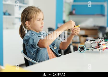 Nettes kleines Mädchen in Casualwear verbindet Details von elektronischen Spielzeug, während am Schreibtisch sitzen bei der Lektion der Robotik und die Schaffung neuer Roboter Stockfoto