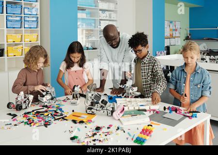 Selbstbewusster Roboterlehrer, der zwischen seinen Schülern steht, Roboter und andere elektronische Spielzeuge konstruiert und ihnen beim Unterricht hilft Stockfoto