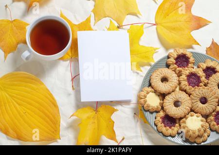 Tasse Tee, verschiedene Kekse auf dem Teller, leeres Papier und Herbstblätter auf weißem Holzhintergrund. Draufsicht mit Textort Stockfoto