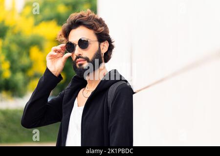 Mann, der eine Sonnenbrille aufsetzt und sich an eine weiße Wand lehnt Stockfoto