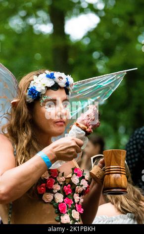Maryland Renaissance Festival, junge Frau im Elfkostüm, aß truthahn Bein und nippte Ale in einem Holzbecher. Girlande aus Blumen und geblümten Weste. Stockfoto