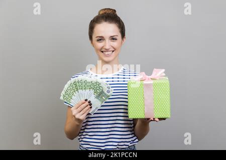 Geschenkeinkäufe. Porträt einer Frau mit gestreiftem T-Shirt, das eine Schachtel mit Geldscheinen hält, und lächelnd vor der Kamera, zufrieden mit dem Kauf. Innenaufnahme des Studios isoliert auf grauem Hintergrund. Stockfoto