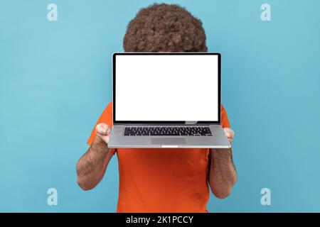 Unbekannter anonymer Mann mit Afro-Frisur, der ein orangefarbenes T-Shirt trägt und sein Gesicht mit einem Laptop mit leerem weißen Display versteckt, Platz für Werbetexte. Innenaufnahme des Studios isoliert auf blauem Hintergrund. Stockfoto