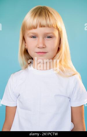 Niedliches Kinderporträt. Frohe Kindheit. Positive Einstellung. Selbstbewusstes, süßes blondes kleines Mädchen in weißem T-Shirt, das die Kamera isoliert auf blauem Backgrou anschaut Stockfoto