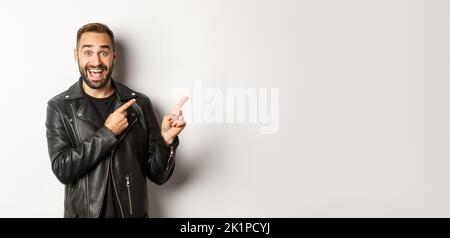 Aufgeregt, schöner Mann in schwarzer Lederjacke, Zeigefinger direkt auf Promo-Angebot, zeigt Logo, weißer Hintergrund Stockfoto