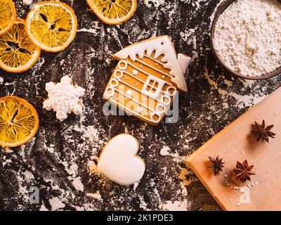 Weihnachtsbäckerei. Traditionelle Kekse. Hausgemachte Küche. Lebkuchenpuderhaus mit Stern- und Herzmehl getrockneten Orangengewürzen auf schwarzem Schreibtisch. Stockfoto
