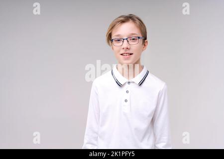 Zuckender kaukasischer Junge, zweifelende zuckende Schultern, Schuljunge, Teenager mit Brille in einem weißen T-Shirt. Der Junge wählt auf einem grauen Hintergrund. Stockfoto