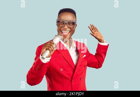 Cooler, exzentrischer und stylischer Showman mit dunkler Haut spricht auf hellblauem Hintergrund ins Mikrofon. Stockfoto