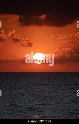 Vertikales Sonnenuntergangsfoto. Die Linie, die das Meer und den Himmel trennt. Gelbe helle Sonnenscheibe hinter Wolken. Übergang von Tag zu Nacht. Stockfoto