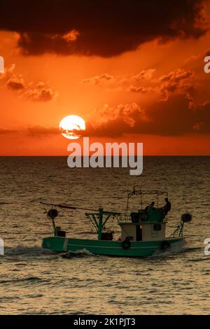 Kleines Fischerboot, das vor der Sonnenuntergangskulisse segelt. Sonnenscheibe hinter Wolken. Farben bei Sonnenuntergang oder Dämmerung. Orangefarbener Himmel. Vertikales Foto. Stockfoto