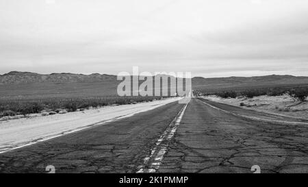 Eine Graustufenaufnahme einer alten, leeren Asphaltstraße, die von einer Landschaft umgeben ist Stockfoto