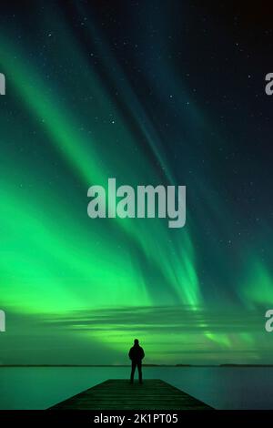 Mann, der auf dem Pier steht und Nordlichter beim Tanzen über den Nachthimmel über der Seenlandschaft beobachtet. Stockfoto