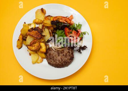 Drei gebratenes Hackfleisch mit gebratener Kartoffel und frischem Salat auf dem Teller auf Gelb Stockfoto