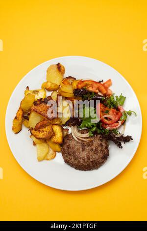 Drei gebratenes Hackfleisch mit gebratener Kartoffel und frischem Salat auf dem Teller auf Gelb Stockfoto