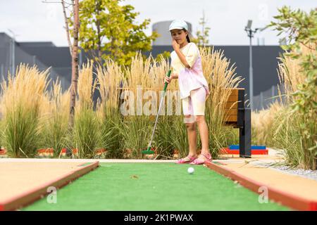 Nettes Mädchen im Vorschulalter, das mit der Familie Minigolf spielt. Glückliches Kind, das Spaß mit Aktivitäten im Freien hat. Sommersport für Kinder und Erwachsene, im Freien Stockfoto