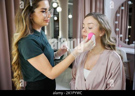 Junge Frau Make-up-Künstler mit Blending Beauty Schwamm während der Anwendung der Grundlage auf dem Gesicht des Kunden. Professionelle Stylistin in sterilen Handschuhen Make-up für Frau im Schönheitssalon. Stockfoto