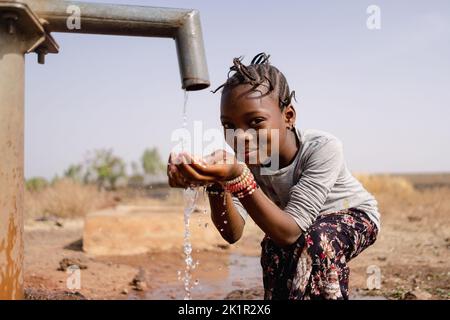 Lächelndes afrikanisches Mädchen, das frisches Wasser aus einem Wasserhahn in einer kargen ländlichen Landschaft mit Landschaft, Zugang zu sauberem Wasser und sanita kostet Stockfoto