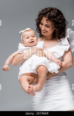 Stilvolle Mama Blick auf lächelnde Mädchen in weißem Kleid und Stirnband isoliert auf grau, Stock Bild Stockfoto