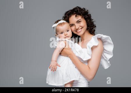 Lächelnde Mutter, die auf die Kamera schaut, während sie das Mädchen in weißen Kleidern hält, isoliert auf grauem, Stockbild Stockfoto