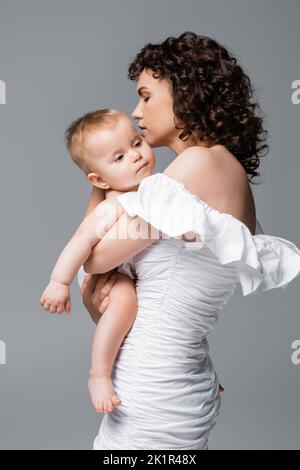 Lockige Frau in Kleid umarmt Säugling Tochter isoliert auf grau, Stock Bild Stockfoto