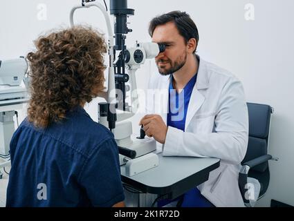 Augenuntersuchung des Kindes in der Augenklinik. Lockiger Junge, der Augenuntersuchung in der Augenklinik mit einem erfahrenen Optiker erhält Stockfoto