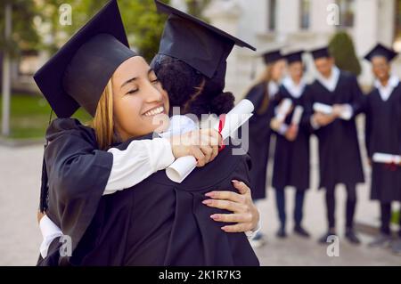 Positive Frauen Absolventen in Studentenkleid umarmen sich nach dem Abschluss von der Universität oder Hochschule Stockfoto