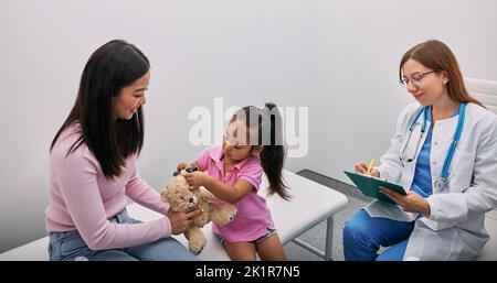 Asiatische Mädchen spielt mit ihrem Spielzeug Bär in medizinischen Spiel, mit Otoskop mit Unterstützung ihrer Mutter und Audiologe. Hörklinik für Kinder Stockfoto