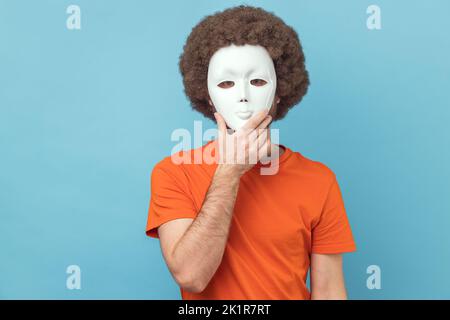 Porträt eines Mannes mit Afro-Frisur, der ein orangefarbenes T-Shirt trägt, das ihr Gesicht mit einer weißen Maske bedeckt und ihre wahre Persönlichkeit, Anonymität, versteckt. Innenaufnahme des Studios isoliert auf blauem Hintergrund. Stockfoto