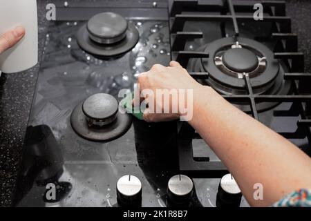 Eine Hausfrau reinigt den Küchengasherd mit einem Schwamm. Wartung von gehärtetem Glas und Glaskeramik. Stockfoto