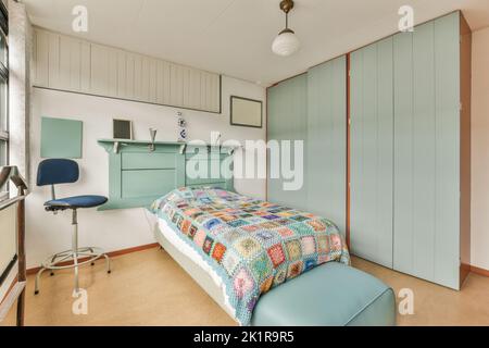 Komfortables Bett mit gepunkteter Federdecke in der Nähe von Kleiderschrank und Tisch mit Stuhl im minimalistischen Stil im modernen Apartment Stockfoto