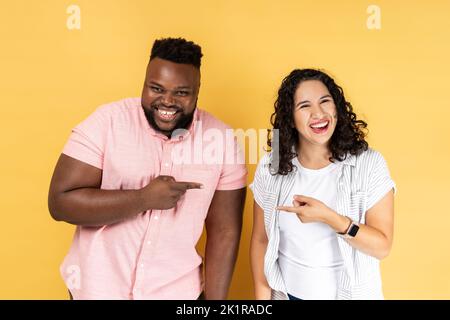 Porträt eines lustigen, positiven jungen Paares in lässiger Kleidung, das zusammen steht, die Finger aufeinander zeigt und lachend auf die Kamera schaut. Innenaufnahme des Studios isoliert auf gelbem Hintergrund. Stockfoto