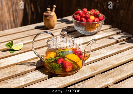 Roter Früchtetee mit Beeren in einer Glas-Teekane auf dem sonnigen Balkon. Stockfoto