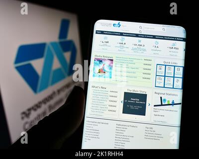 Person, die Mobiltelefon mit einer Webseite des israelischen Zentralbüros für Statistik (CBS) auf dem Bildschirm vor dem Logo hält. Konzentrieren Sie sich auf die Mitte des Telefondisplays. Stockfoto