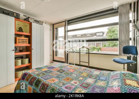 Komfortables Bett mit gepunkteter Federdecke in der Nähe von Kleiderschrank und Tisch mit Stuhl im minimalistischen Stil im modernen Apartment Stockfoto