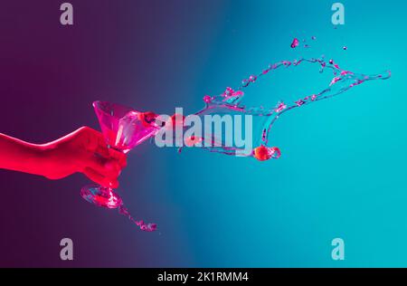 Weibliche Hand, die über den Martini-Cocktail spritzt, spritzt auf blauem Hintergrund in pinkem Neonlicht. Nachtleben, Party. Stockfoto