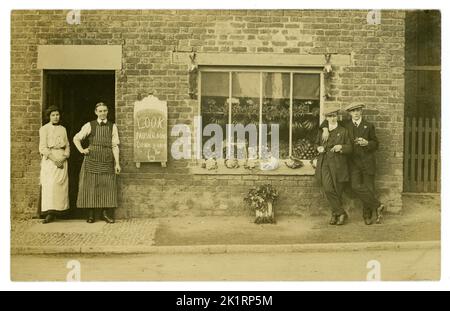 Originale, sehr klare Postkarte aus der Zeit der frühen 1900er Jahre mit einem kleinen Lebensmittelgeschäft, einem Gemischtwarenladen, einem unabhängigen Geschäft, der Gemüse verkauft - Blumenkohl, Sellerie, Obst - Bananen -, die im Schaufenster ausgestellt wurde. Display Board Werbung Pilze im Preis reduziert. Ein Kaninchen hängt an jeder Seite des Fensters. Ehemann und Ehefrau, Paar, Besitzer (der Mann mit Streifenschürze) stehen vor ihrem Laden mit 2 modischen Jungs in Anzügen und flachen Mützen, Kunden, frechen Charakteren, Freunden, Freundschaften, vielleicht schwul. Edwardianisches Geschäft / viktorianisches Geschäft. Um 1912. Dorf in Großbritannien Stockfoto