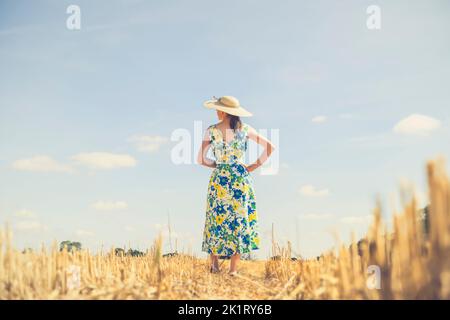 Rückansicht einer Frau in einem Sommerkleid und Strohhut auf einem Feld mit blauem Himmel an einem sonnigen Tag. Stockfoto