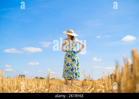 Rückansicht einer Frau mit Blumenkleid und Strohhut, die an einem sonnigen Tag auf einem ausgeernteten Feld mit blauem Himmel steht. Stockfoto