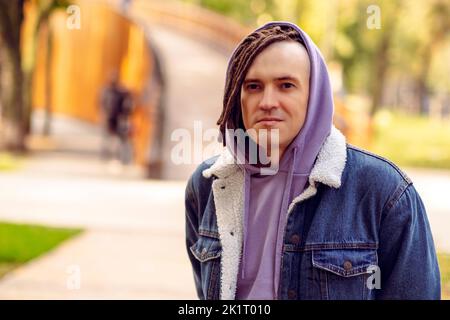Porträt eines gutaussehenden Mannes mit braunen Dreadlocks im Hoodie, Denim-Jacke, die auf der Straße im Stadtpark posiert Stockfoto