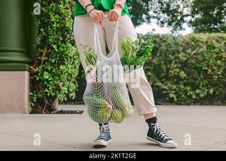 Nicht erkennbare Frau in grünem Hemd, die eine Netztasche mit Gemüse hält, auf der Straße stehend, Nahaufnahme Stockfoto